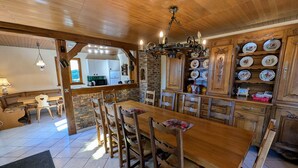 A wooden dining table with chairs, a chandelier, and a kitchen area with cabinets and a brick wall.