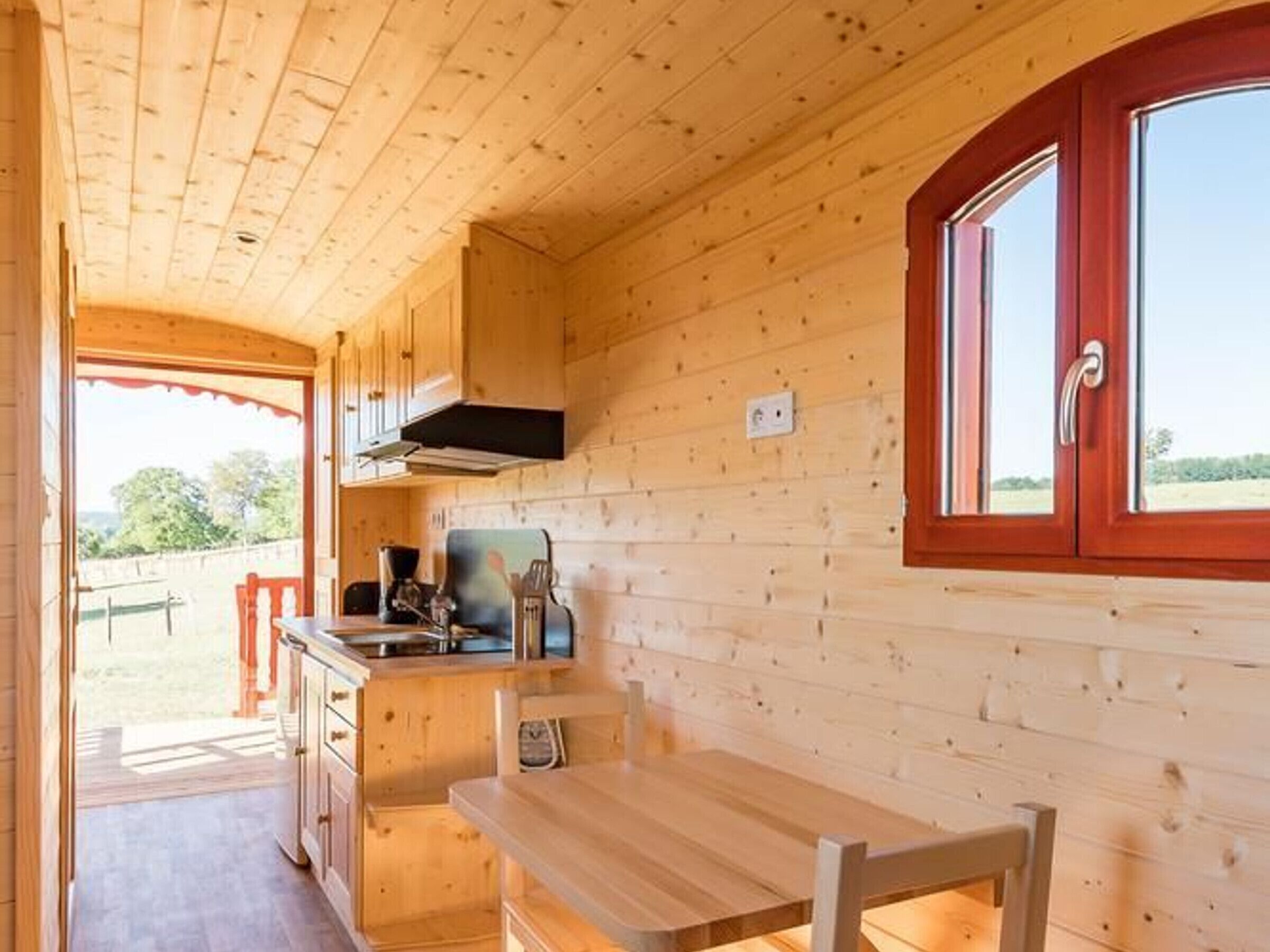 A wooden cabin interior with a kitchen area, a dining table, and a window with red trim.