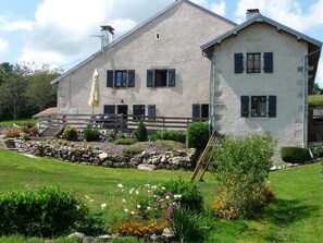 A large, two-story stone building with a sloped roof, a wooden deck with outdoor seating, and a well-maintained garden with various flowers and shrubs.