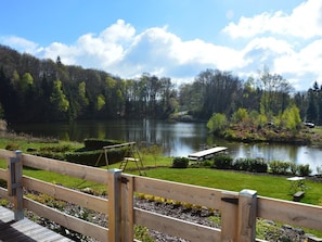 A wooden deck with a view of a lake, surrounded by trees and a grassy area with a bench and a small table.