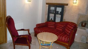 Una sala de estar acogedora con un sofá rojo, una silla de madera y una pequeña mesa redonda. Hay dos lámparas fijadas a la pared y una ventana con vistas al exterior.