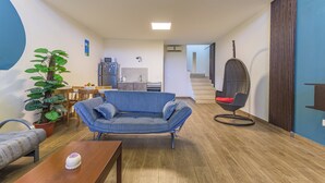 A living room with a sofa, a dining table, and a hanging chair. There is a kitchen area in the background with a refrigerator and a stove. - Mavi 1BR Apartment With Jacuzzi In Batroun (Batroun)