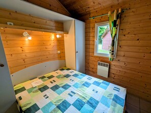 A small, cozy room with a wooden ceiling, a single bed with a checkered quilt, a window with curtains, and a wall-mounted heater.