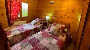 En hytte med to senge, trævægge, et vindue med gardiner og et lille bord med en lampe.