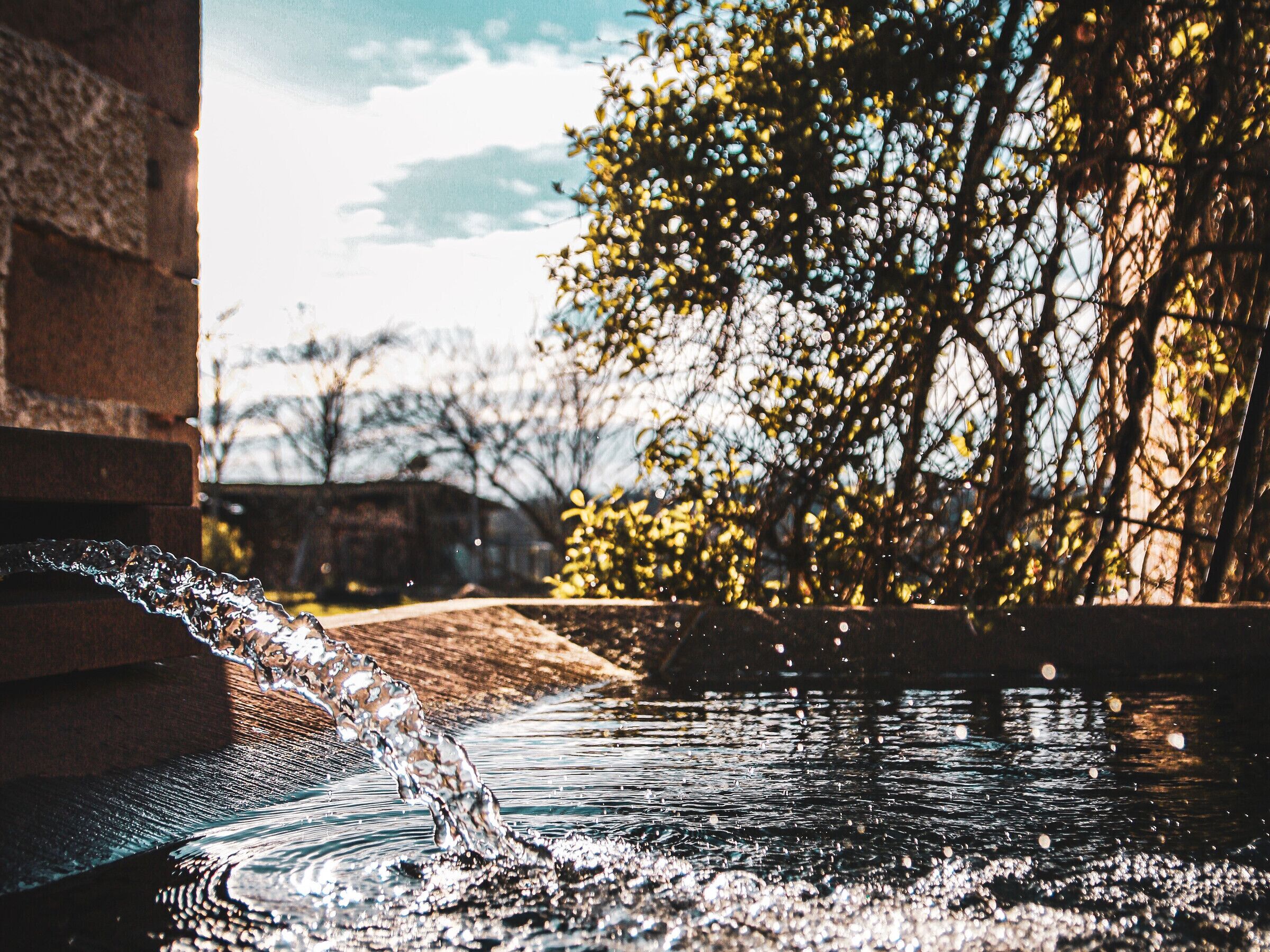 Un élément décoratif aquatique composé d’un ruisseau aux eaux claires se jetant dans un bassin aux reflets changeants, le tout entouré d’arbres et d’une structure en briques.