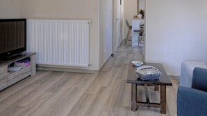 Un salon équipé d’un téléviseur, d’une table sur laquelle se trouvent divers objets, d’un radiateur, et d’un couloir menant à une salle à manger.
