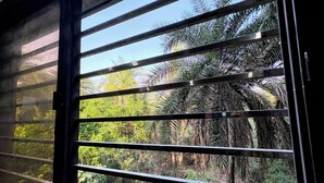 A window with horizontal bars, offering a view of dense greenery and palm trees outside. - sukoon homestay (Agra)