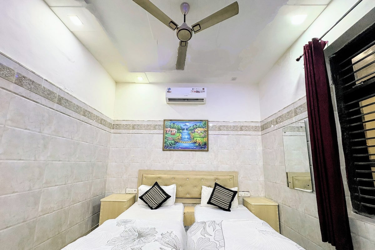 A hotel room with two beds, a ceiling fan, an air conditioning unit, a painting on the wall, and a window with curtains.