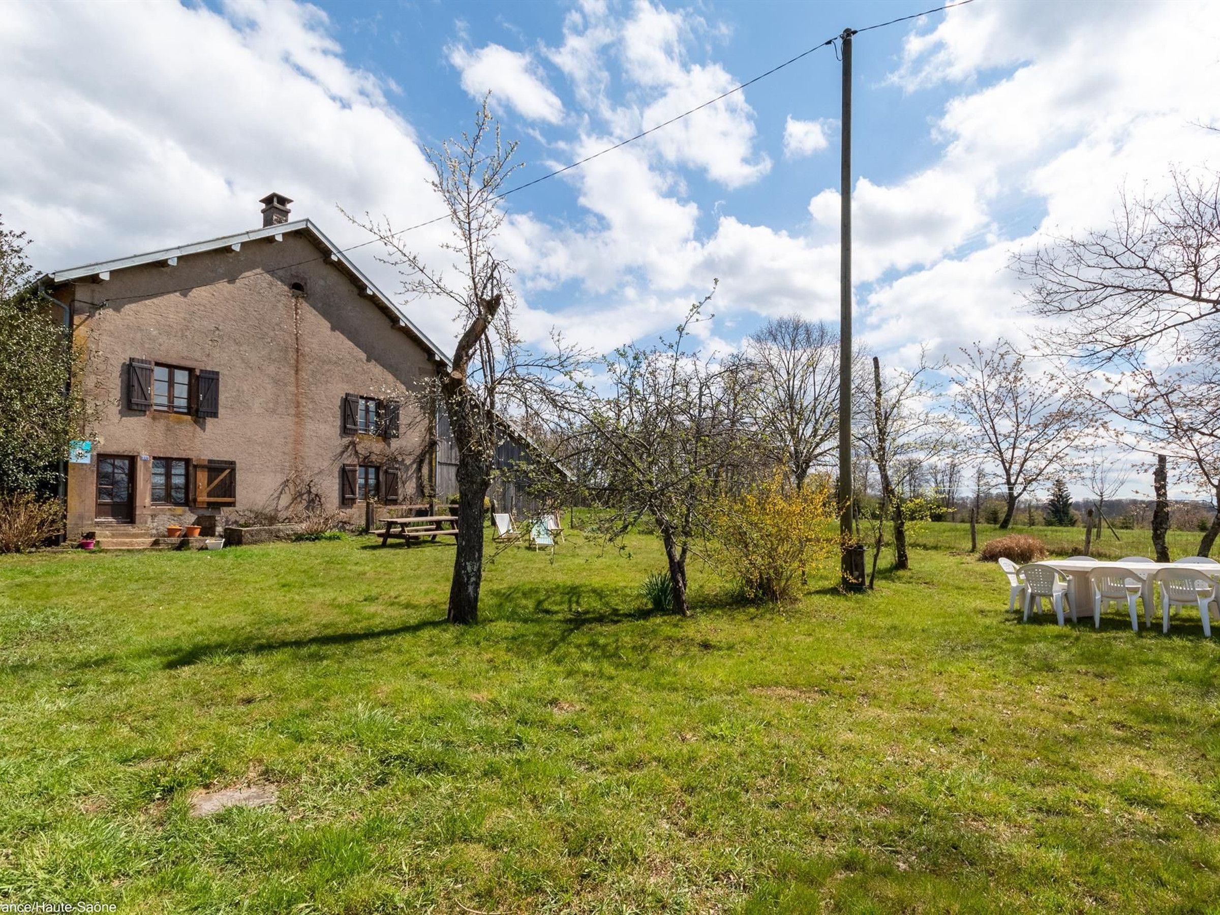 A rustic house with a grassy yard, a picnic table, and a row of white plastic chairs.