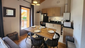A compact kitchen with wooden cabinets, a black refrigerator, and a microwave. There is a dining table with chairs and a window providing natural light.