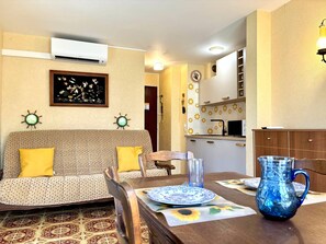 A living room with a sofa, a dining table with a pitcher, and a kitchen area with patterned tiles.