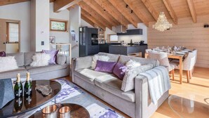 A spacious living area with a high ceiling and wooden beams, a cozy seating arrangement, and a modern kitchen in the background. - Cozy chalet in Méribel with jacuzzi, sauna, 4 bedrooms (Méribel)