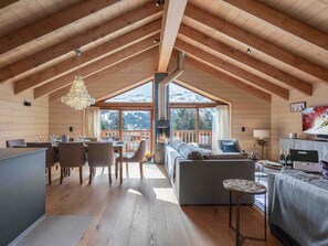 Een ruime woonkamer met een hoge plafond, houten balken, een open haard en een groot raam met uitzicht op een besneeuwde berg.
