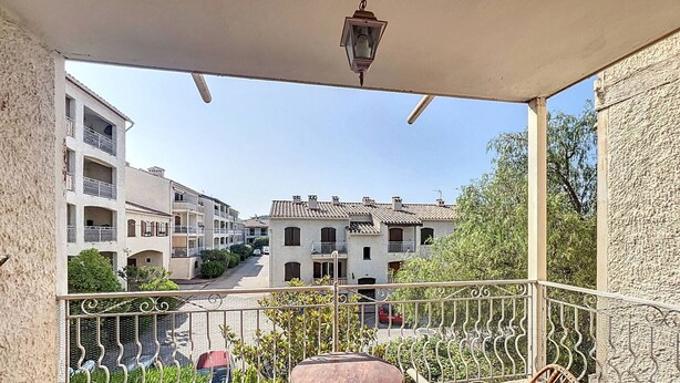 Un balcon avec des fauteuils en osier et une table, donnant sur une rue bordée d’immeubles et de verdure.