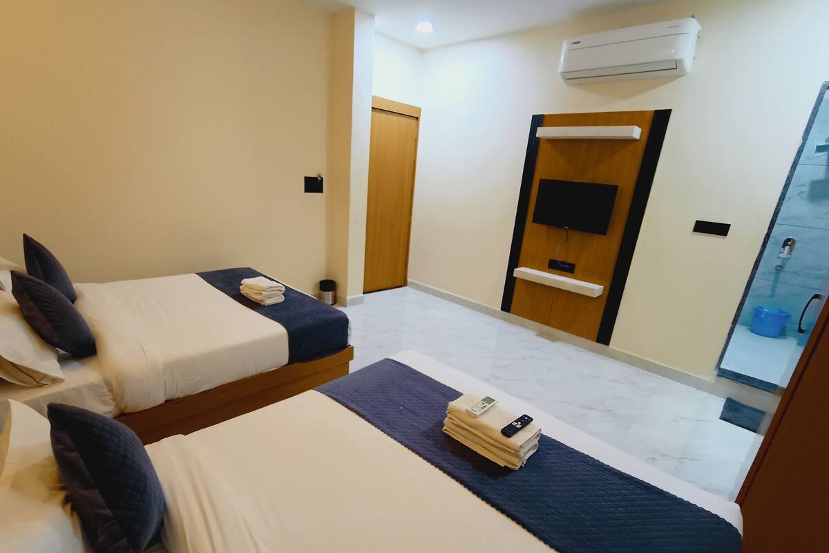 A hotel room with two beds, a TV, an air conditioner, and a bathroom visible through a glass door.
