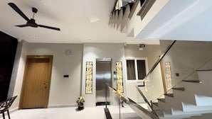 Egy modern belső tér, melyben van egy fa ajtó, egy mennyezeti ventilátor, egy virágtartóban lévő növény, egy üveg lépcsőház és egy rozsdamentes acélból készült lift.