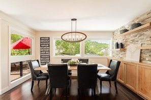 A dining area with a wooden table, black chairs, a chandelier, and a view of greenery through large windows. - Mountain View Deer Valley Escape | Hot Tub | Near Hiking & Biking Trails (Park City)