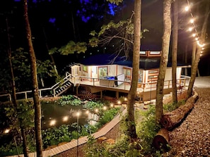 A houseboat with a deck, stairs, and a small dock extending into a body of water, surrounded by trees. - Tranquil Hideaway - Romantic Spa Retreat with Hot Tub and Sauna (Denniston)