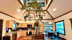 A modern kitchen with a central island, a dining area with a large window, and a staircase leading to an upper level. - Tranquil Hideaway | Romantic Spa Retreat with Hot Tub, Cold Plunge and Sauna (Denniston)