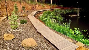 A wooden pathway leading to a pond with string lights, surrounded by rocks and greenery. - Tranquil Hideaway | Romantic Spa Retreat with Hot Tub, Cold Plunge and Sauna (Denniston)