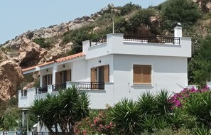 Un bâtiment blanc doté d’un balcon, entouré de verdure et adossé à une colline rocheuse.