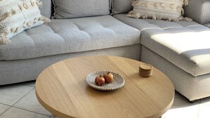 Een grijze modulaire bank met een geweven plaid en bijpassende kussens, een ronde houten salontafel met een geweven mand en granaatappel, en een stapel houten onderzetters.