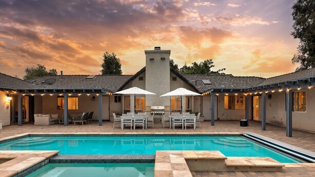 A swimming pool with a tiled deck, outdoor seating area, and a building with a tower-like structure in the background.