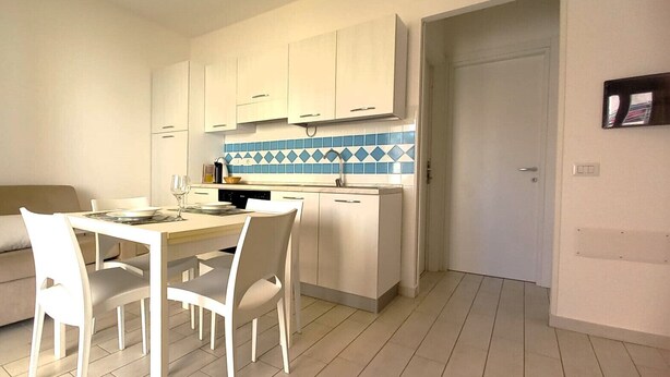 Una cocina compacta con armarios blancos y salpicadero de azulejos azules, una mesa de comedor con sillas y un sofá beige en el área de estar contigua.