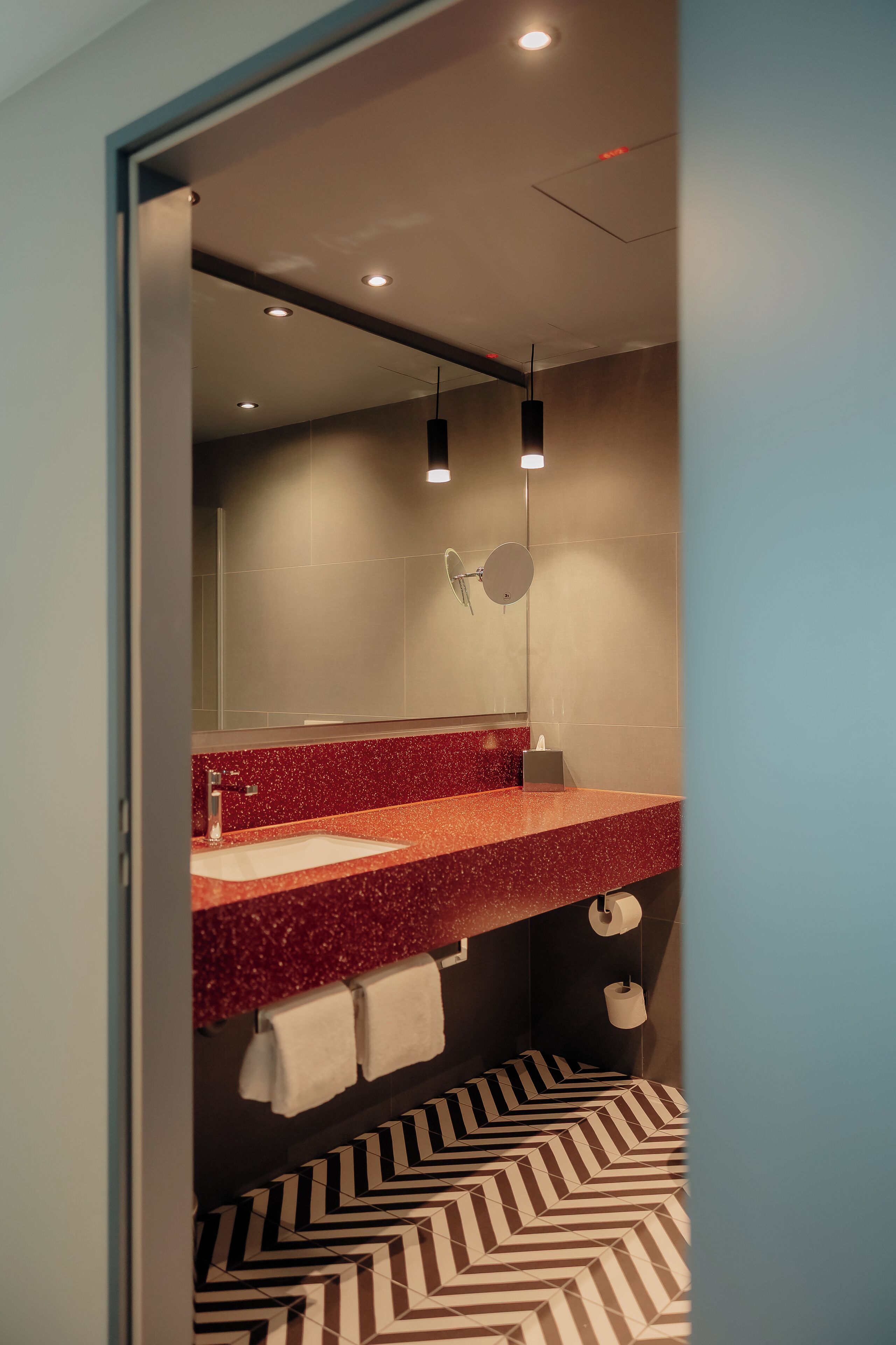 Ein modernes Badezimmer mit einer roten Arbeitsplatte mit Speckmuster, einem großen Spiegel und einem Fußboden mit Muster.