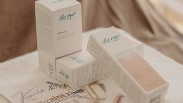 Mehrere La Mer Serum-Verpackungen auf einem weißen Handtuch, zusammen mit einem Zertifikat und einer kleinen Pflanze.