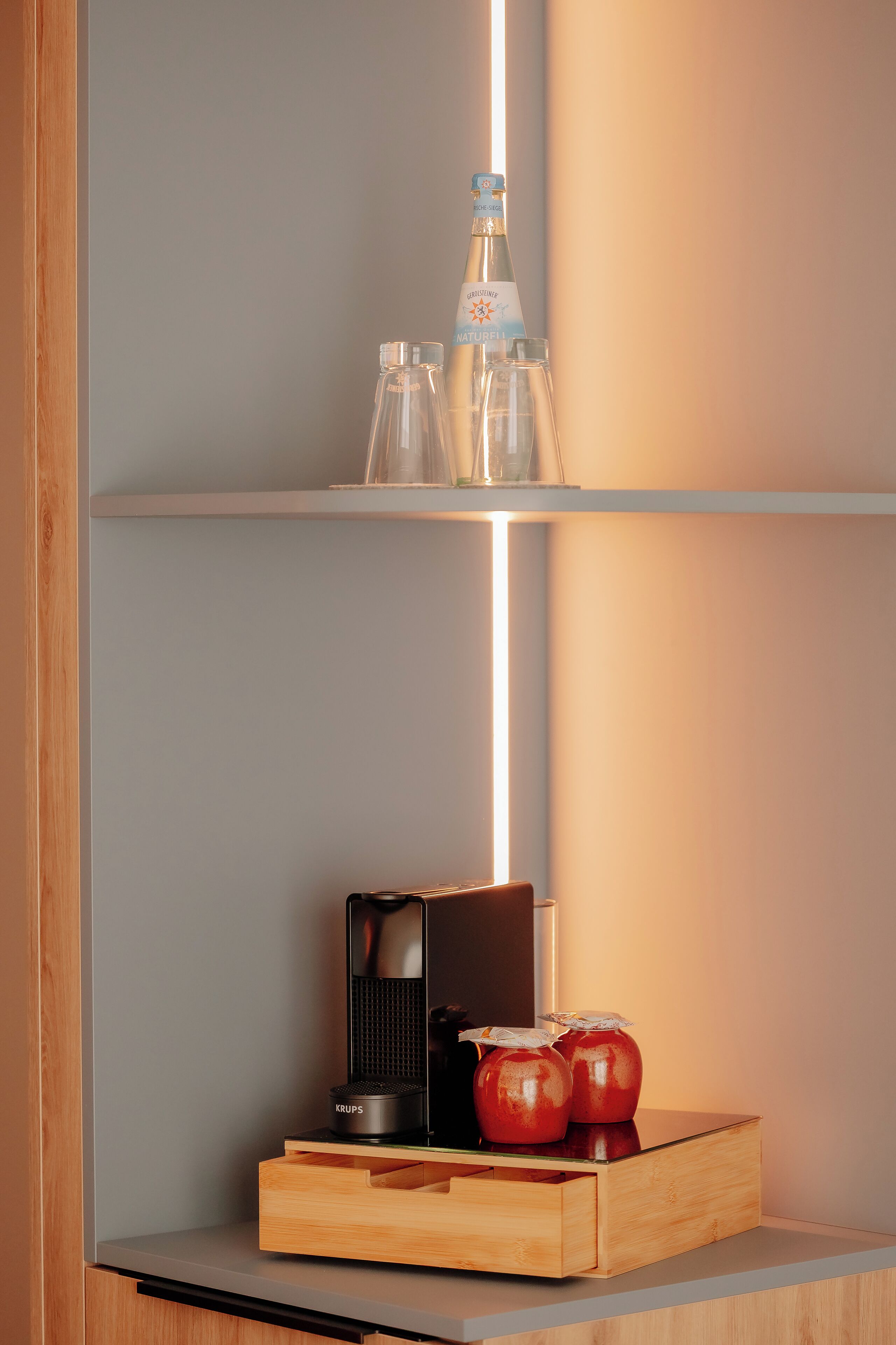 A kitchen with a built-in microwave, a wooden drawer, and a shelf with a bottle and two glasses.