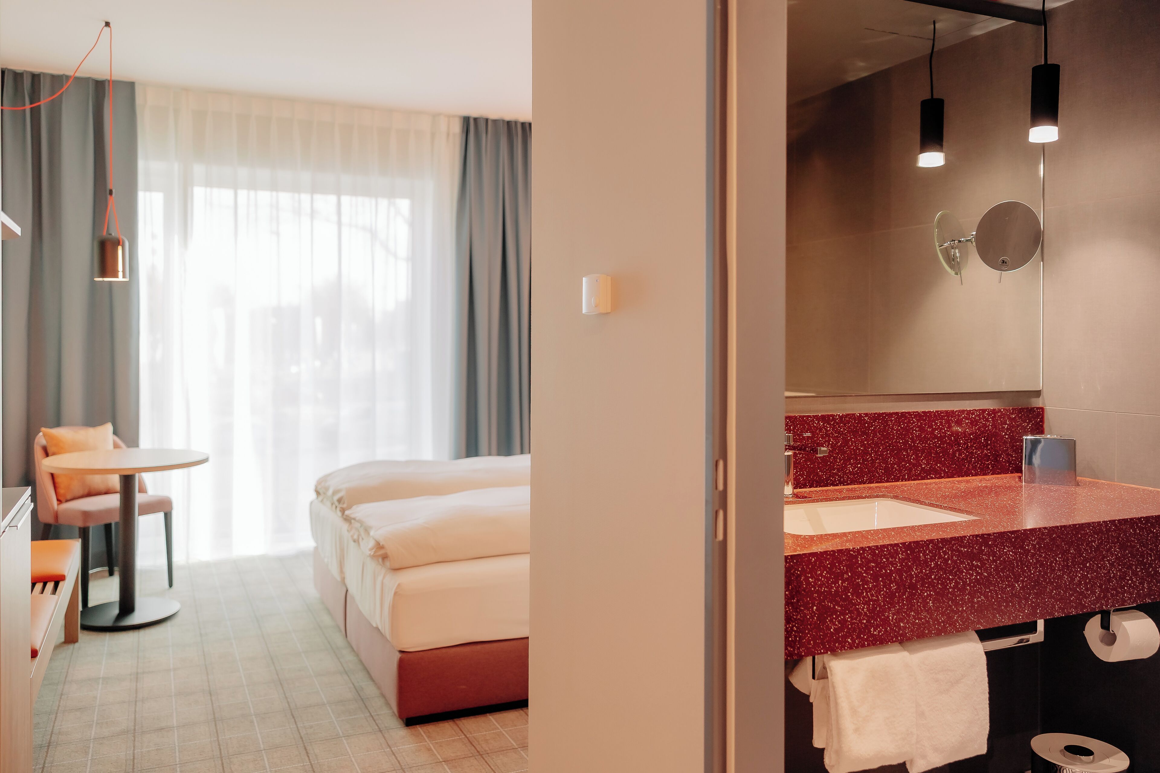 A hotel room with a bed, a desk with a chair, a round table, a bathroom with a red countertop, and a mirror.