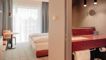 A hotel room with a bed, a desk with a chair, a round table, a bathroom with a red countertop, and a mirror.