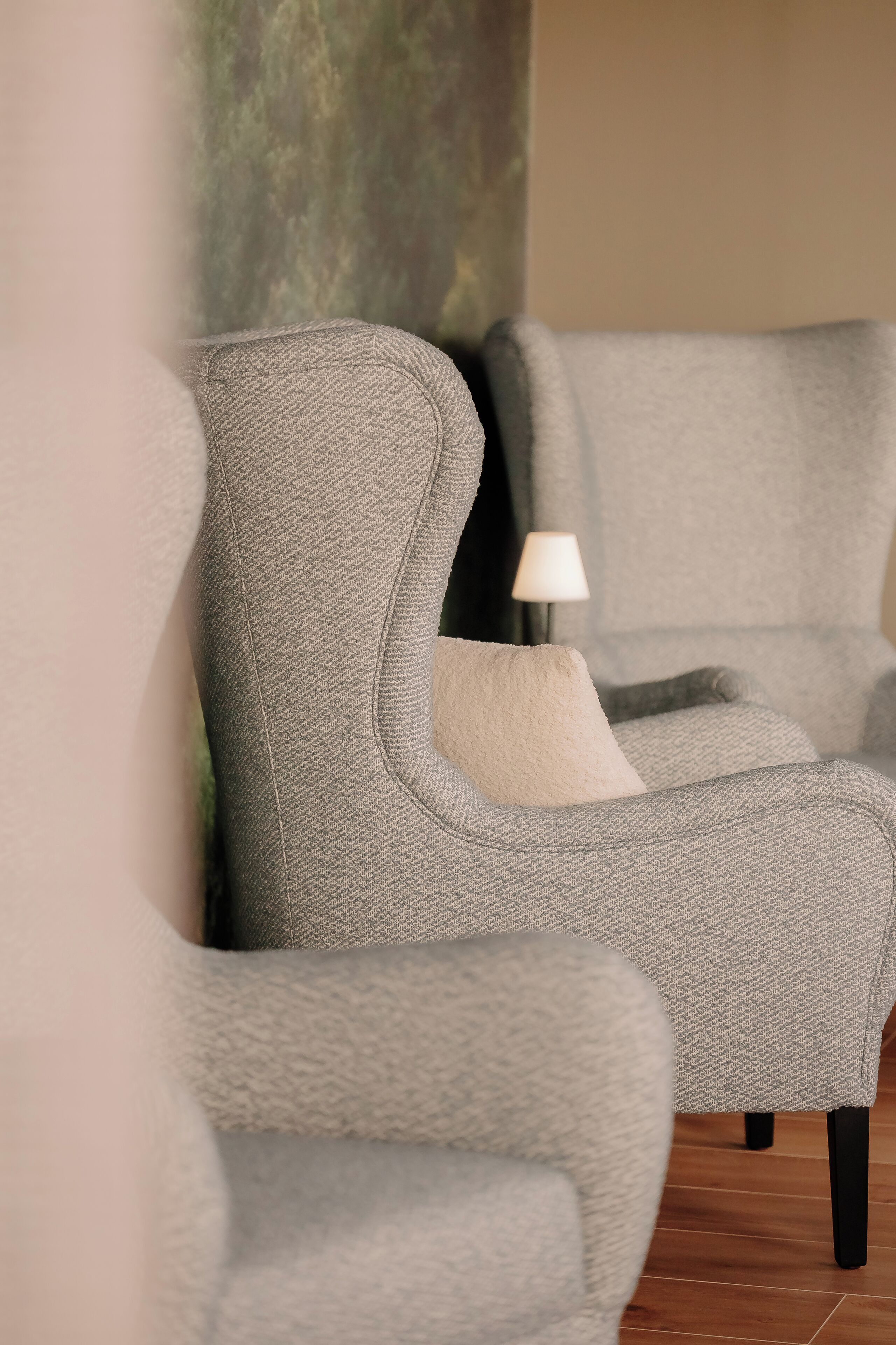 Een grijze bekleden fauteuil met een kussen, geplaatst tegen een muur met een structuur.