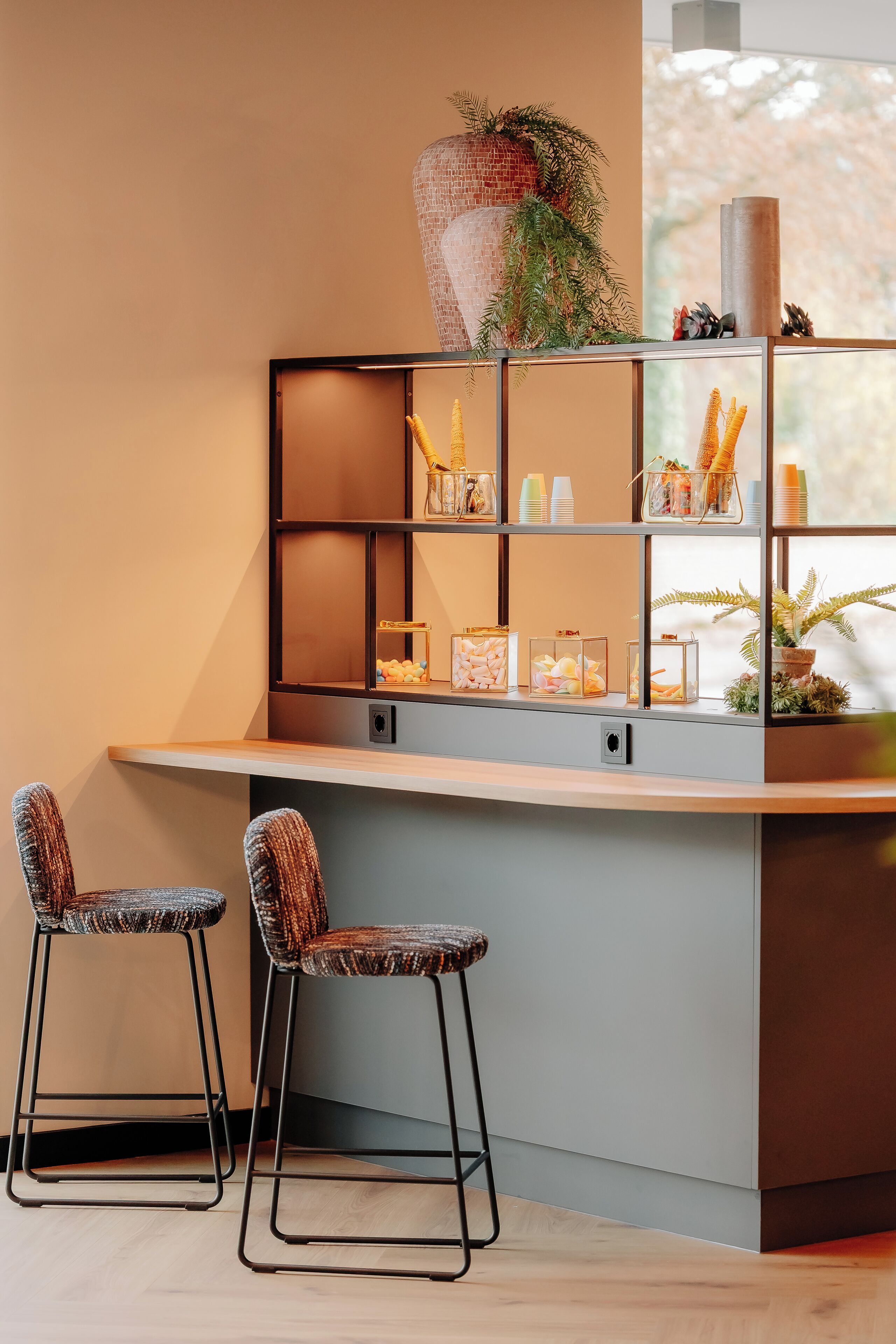 Un área de bar moderna con dos taburetes, una estantería con diversos objetos y una planta decorativa.