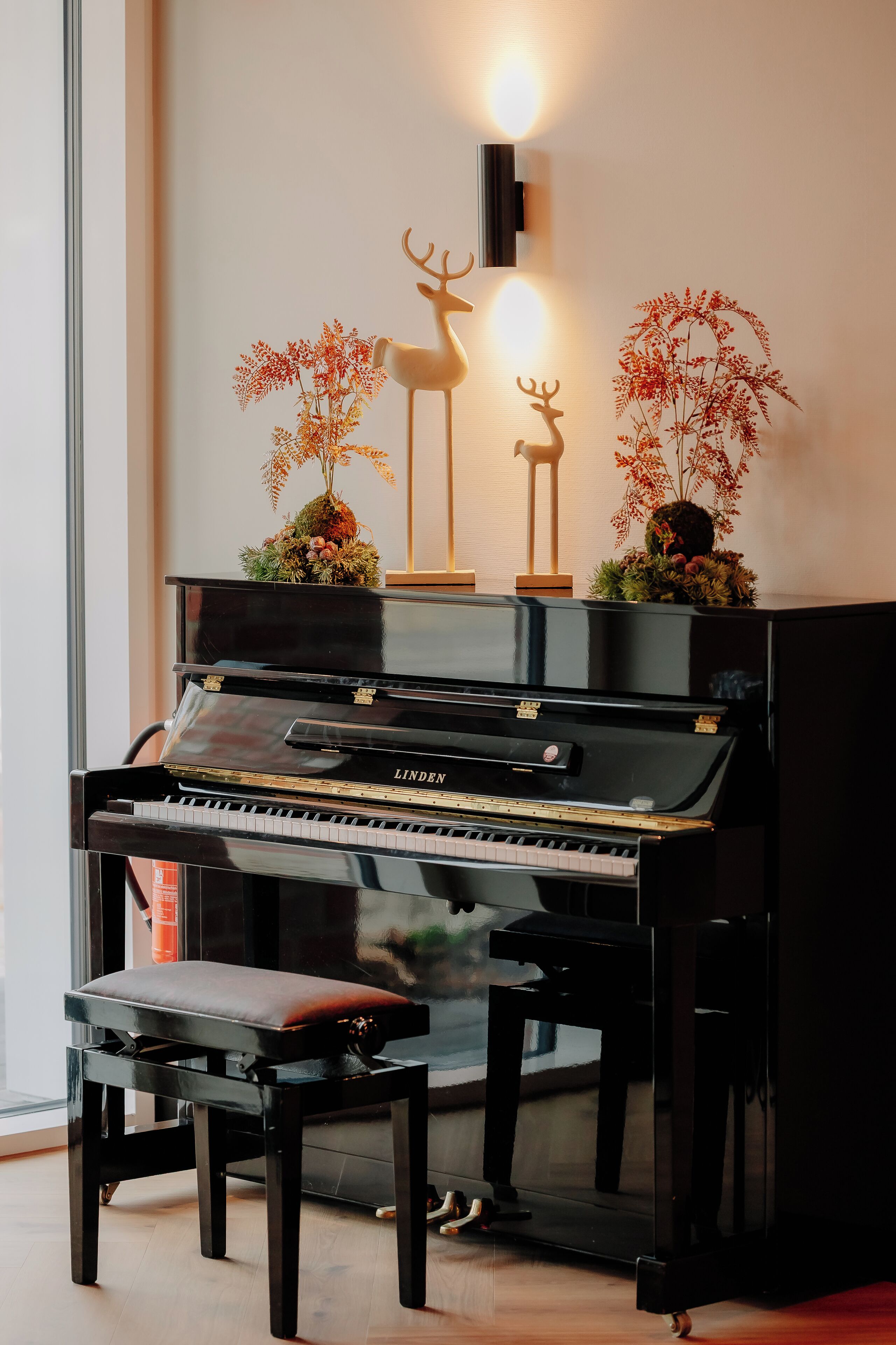 Un piano negro con un banco, una lámpara fijada a la pared y dos figuras decorativas de ciervos con plantas.