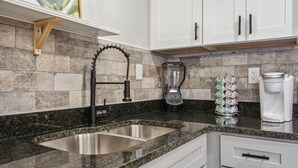 A kitchen with a granite countertop, stainless steel sink, a blender, a coffee maker, and a stack of coffee pods.
