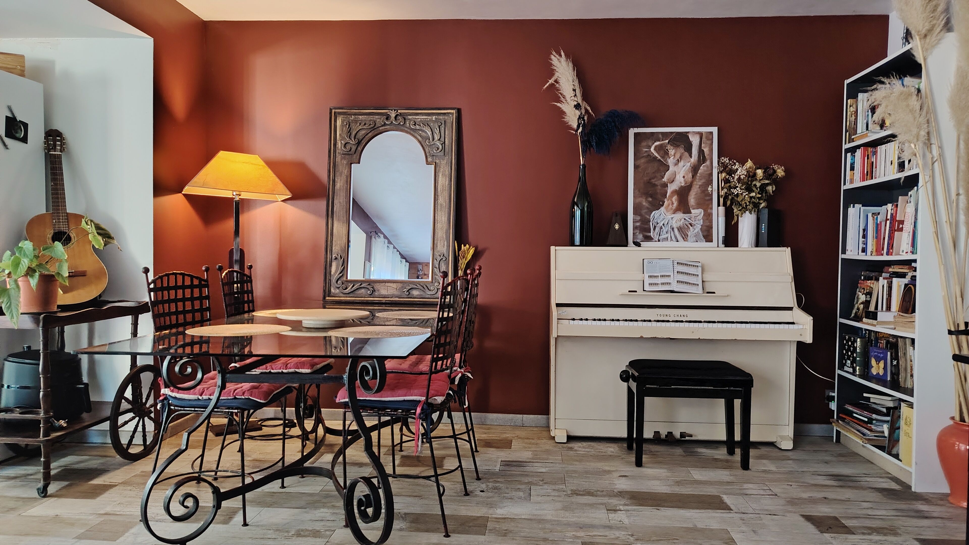 A room with a piano, a table with chairs, a guitar, a lamp, and a bookshelf.