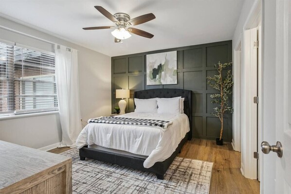 Une chambre à coucher avec un lit, un ventilateur de plafond, une fenêtre avec des stores, une table de chevet et une plante en pot.