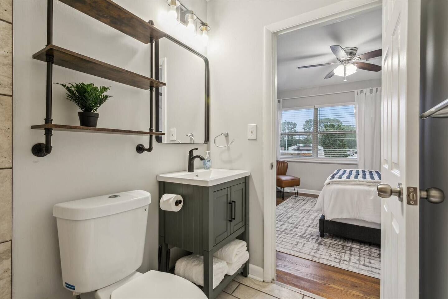 A bathroom with a toilet, sink, and mirror. There is a window with blinds and a view of the outside.