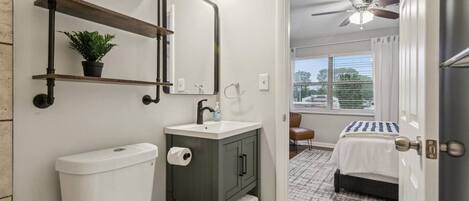A bathroom with a toilet, sink, and mirror. There is a window with blinds and a view of the outside.