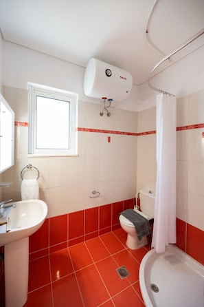 A bathroom with a white sink, toilet, and bathtub, red-tiled floor, and a window above the sink. - Casa Vardani 3 Modern Central Apartment (Lefkada)