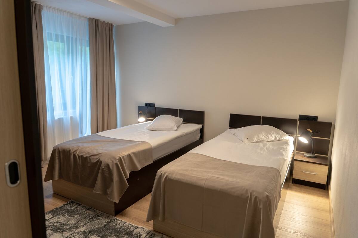 A hotel room with two beds, a nightstand with a lamp, a window with curtains, and a door.