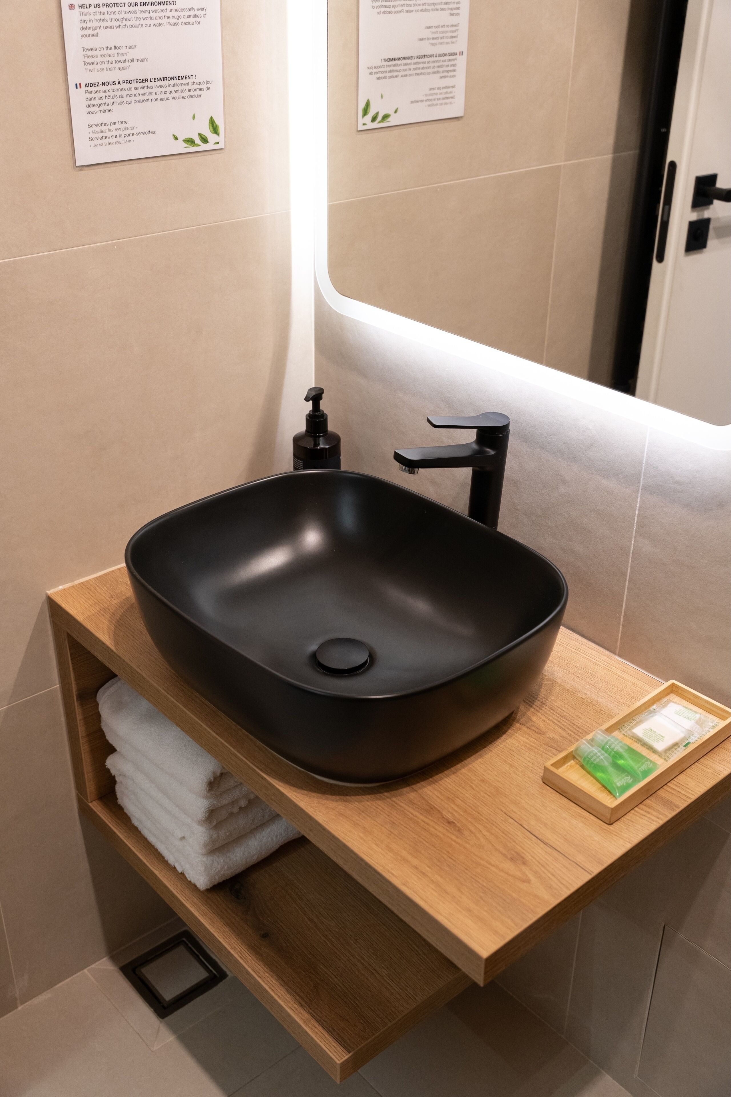 A modern bathroom with a black vessel sink, wooden shelf with towels, and a hand soap dispenser.