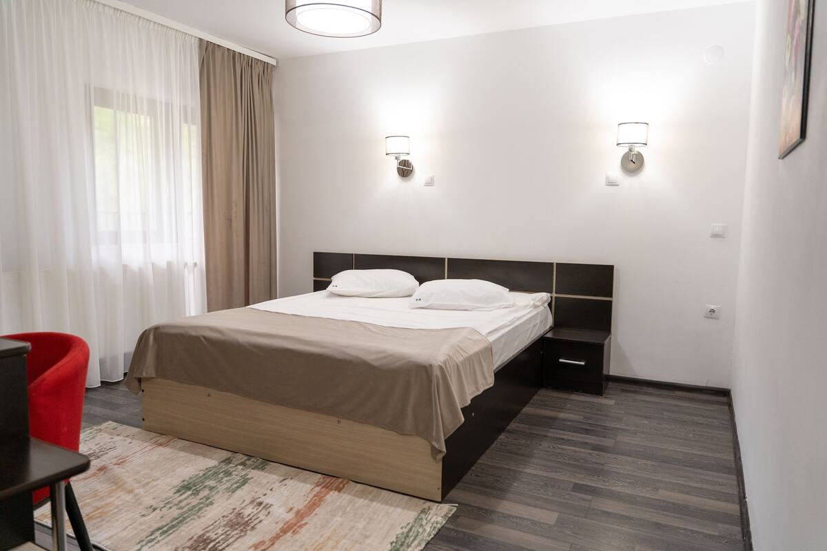 A modern hotel room with a bed, bedside tables, a red chair, and a window with curtains.