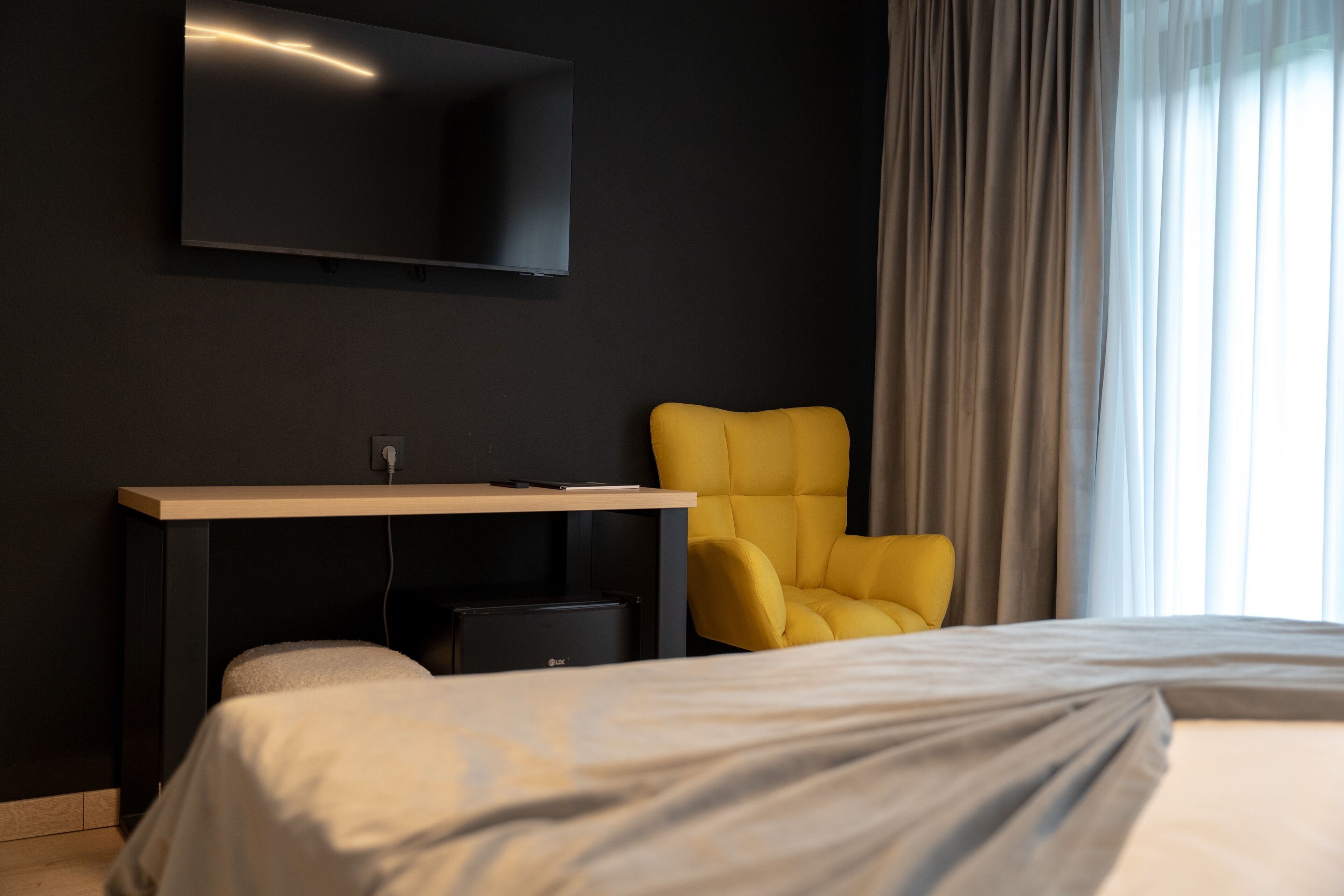 A hotel room with a bed, a desk with a flat-screen TV, a yellow chair, and a large window with curtains.