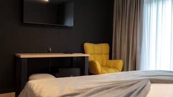 A hotel room with a bed, a desk with a flat-screen TV, a yellow chair, and a large window with curtains.