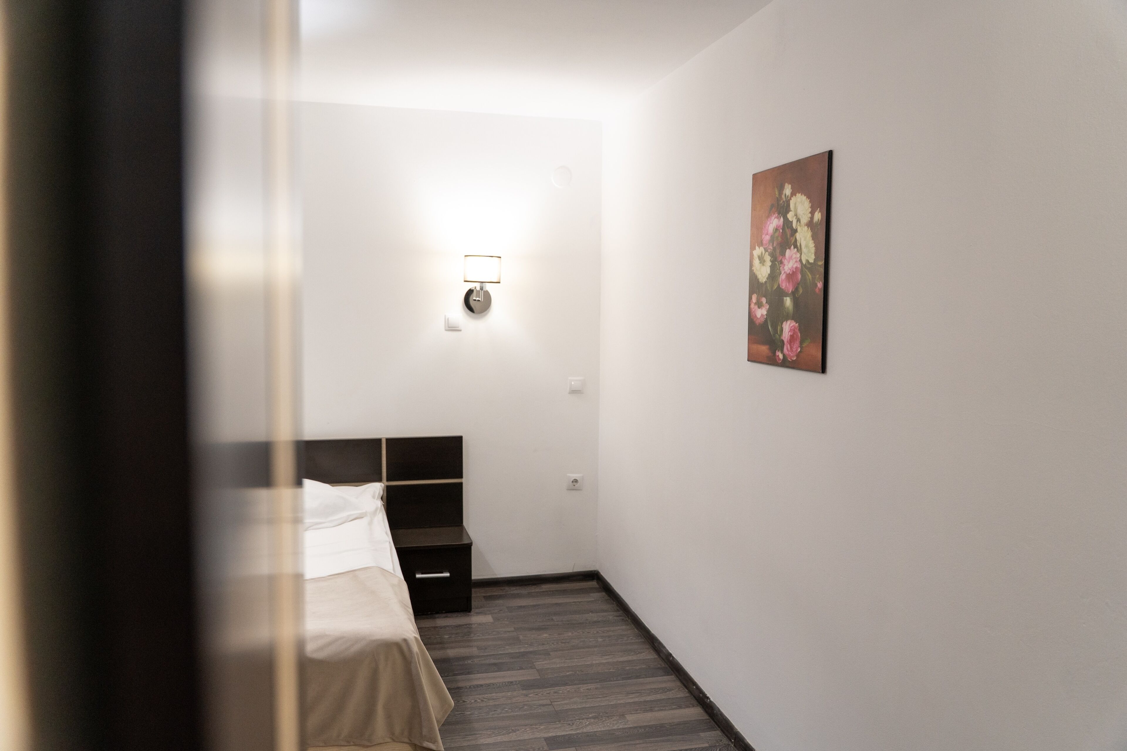A hotel room with a bed, bedside table, wall-mounted lamp, and a floral painting.