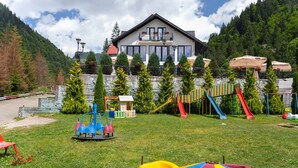 A playground with slides and swings, a building with a balcony, and a mountainous backdrop.