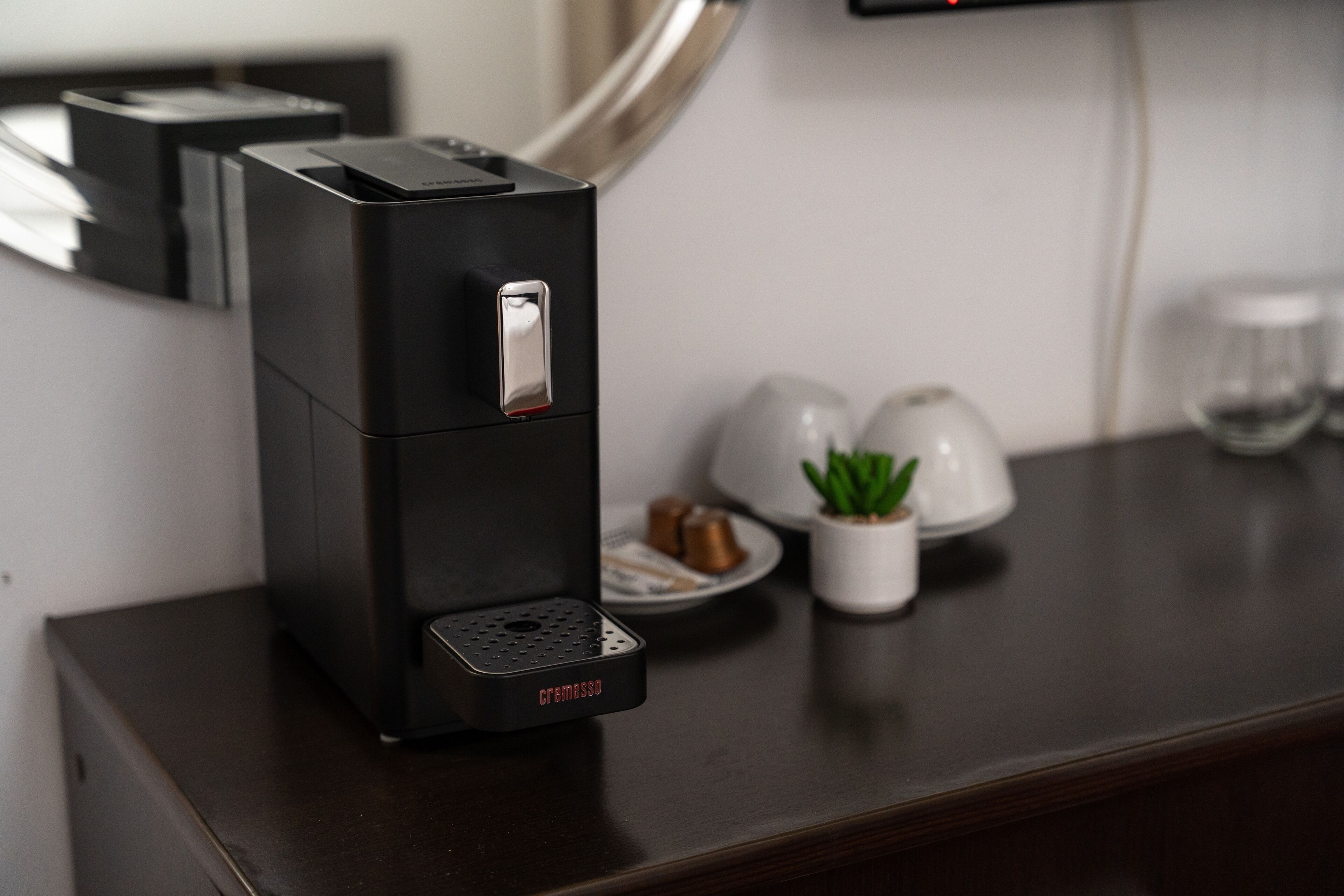 A coffee machine on a dark wooden table with a small potted plant and a glass of water nearby.